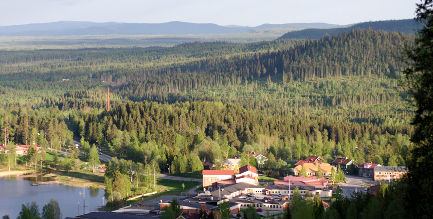vy över samhället, hus, skog och en sjö. I bakgrunden syns berg och himmel.