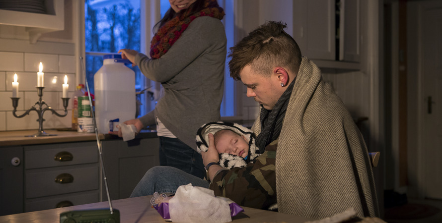 Strömavbrott, två vuxna och en bebis i varma kläder. Levande ljus brinner och en batteriradio står på bordet. 