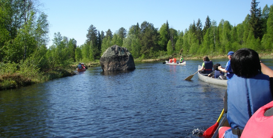 Människor sittandes i kanoter paddlandes på vatten. I bakgrunden syns gröna träd och blå himmel.